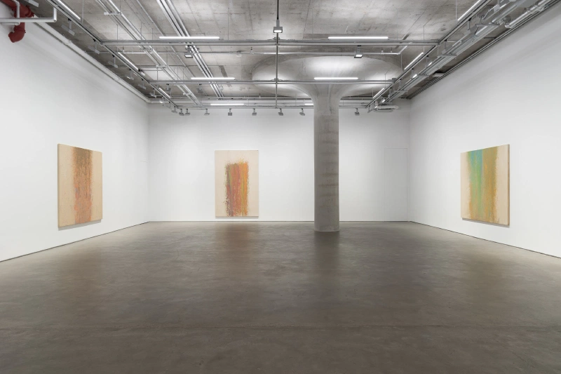 Four pale vertical color-field paintings displayed around a concrete column in a gallery.