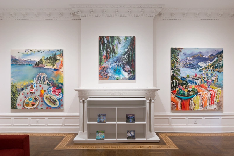 Three landscape and still-life paintings arranged around a fireplace mantel in a gallery.
