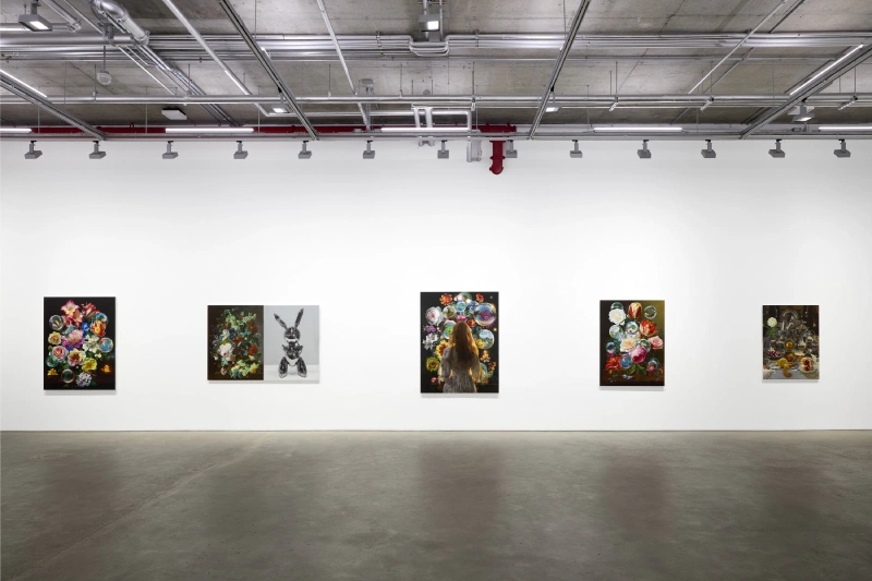 Installation view of hyperrealist still-life paintings displayed across a wide white gallery wall.