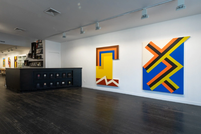 Gallery installation with two geometric paintings and a counter displaying smaller works.