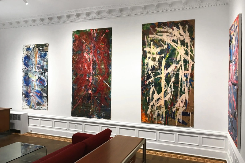 Four large gestural abstract paintings displayed above red seating in a gallery.
