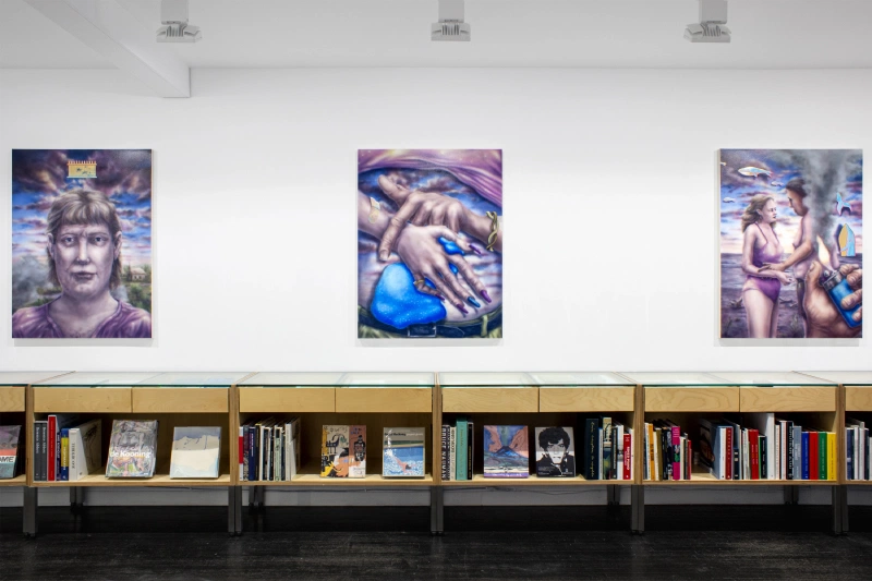 Three figurative paintings displayed above a long bookshelf in a gallery.