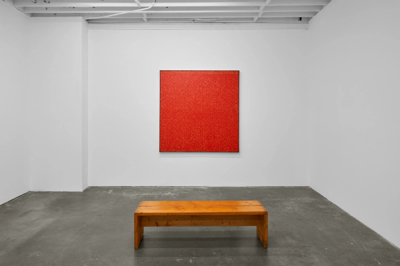 A single red square painting displayed above a wooden bench in a white gallery.